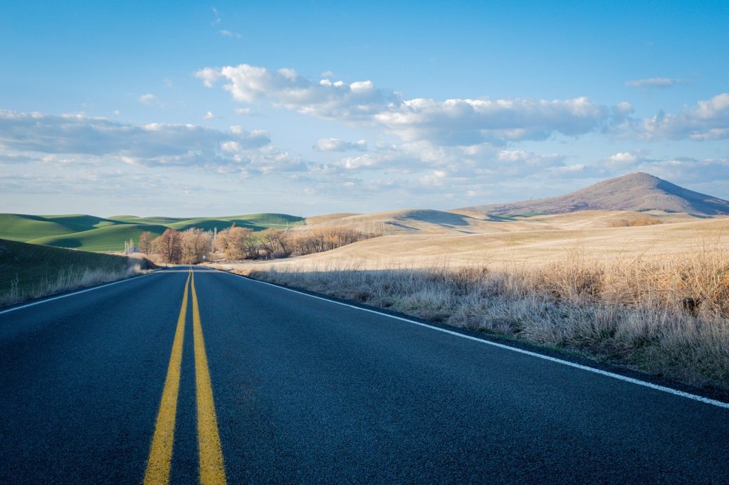 A scenic landscape features a long, empty paved road with double yellow lines stretching into the distance under a bright blue sky with scattered white clouds. On either side of the road, the terrain consists of rolling hills, with some appearing green and others covered in dry, golden grass. In the far distance on the right, a prominent, conical hill or small mountain rises against the horizon. The overall impression is one of open spaces and a journey through a rural or semi-arid environment.