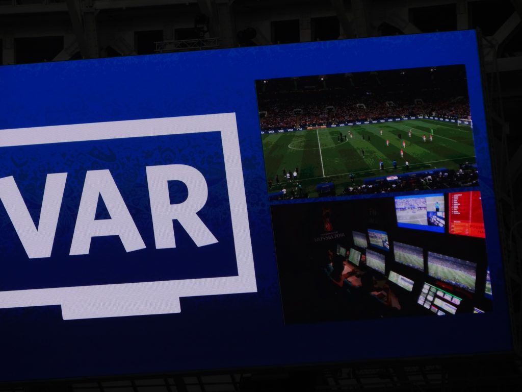 A VAR screen showing a live football match and the control room, representing a technological revolution in Premier League history.