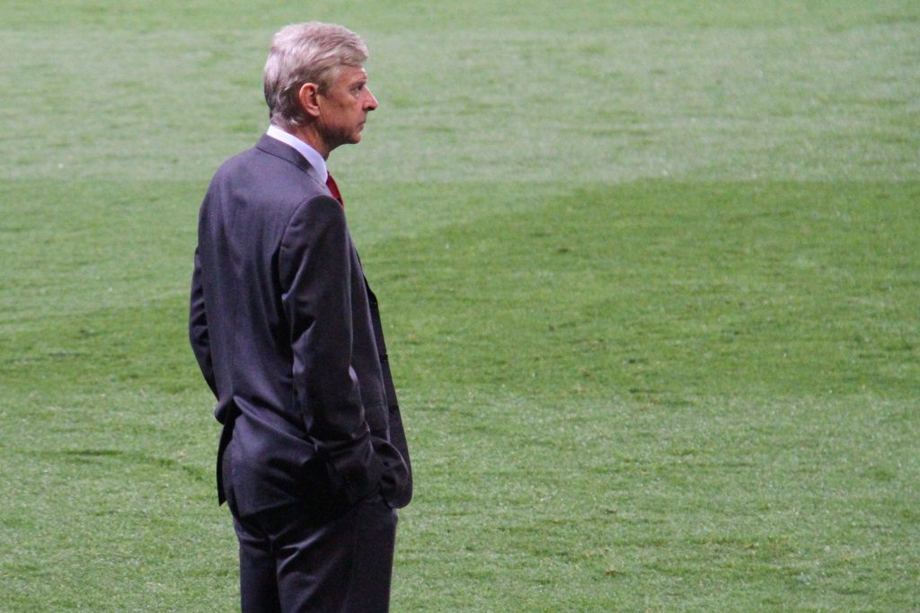 Arsène Wenger with light-colored hair, wearing a dark suit and a red tie, stands on a green football pitch, looking to his right.