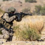 A soldier in full combat gear kneels in a desert-like environment, aiming a rifle. The soldier is wearing camouflage fatigues, a helmet, and a tactical vest, with a pistol holstered on their right hip. The rifle has an optical sight mounted on top. The background consists of dry, light-colored earth and sparse, dry brush, suggesting an arid or semi-arid region.