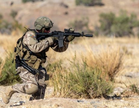 A soldier in full combat gear kneels in a desert-like environment, aiming a rifle. The soldier is wearing camouflage fatigues, a helmet, and a tactical vest, with a pistol holstered on their right hip. The rifle has an optical sight mounted on top. The background consists of dry, light-colored earth and sparse, dry brush, suggesting an arid or semi-arid region.