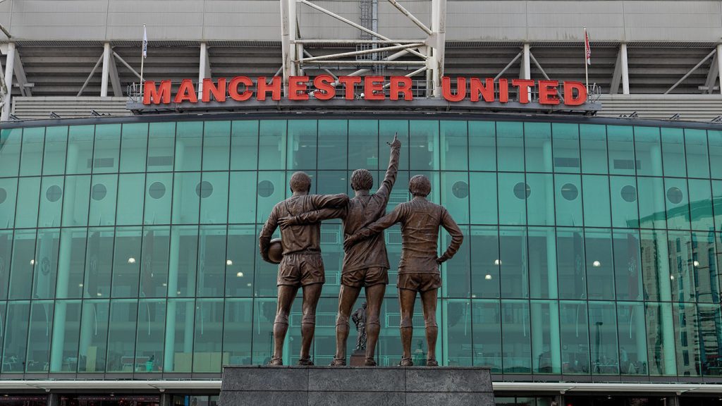 A statue of three Manchester United football players stands in front of Old Trafford, the Manchester United stadium. The players, seen from behind, are embraced, with the middle player pointing upwards. Above the glass facade of the stadium, "MANCHESTER UNITED" is written in red letters.