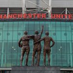 A statue of three Manchester United football players stands in front of Old Trafford, the Manchester United stadium. The players, seen from behind, are embraced, with the middle player pointing upwards. Above the glass facade of the stadium, "MANCHESTER UNITED" is written in red letters.
