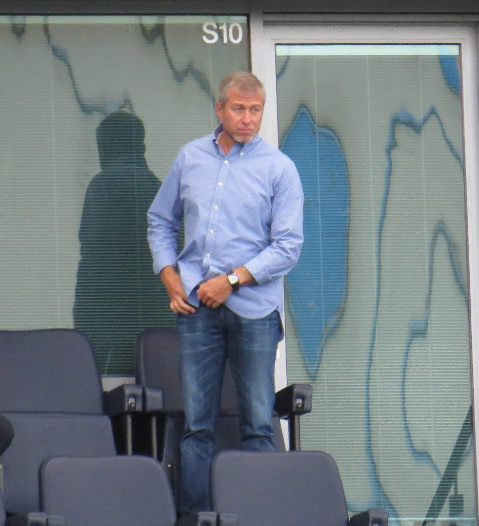Roman Abramovich in a light blue button-down shirt and jeans stands in what appears to be a stadium box or lounge, with "S10" visible above.