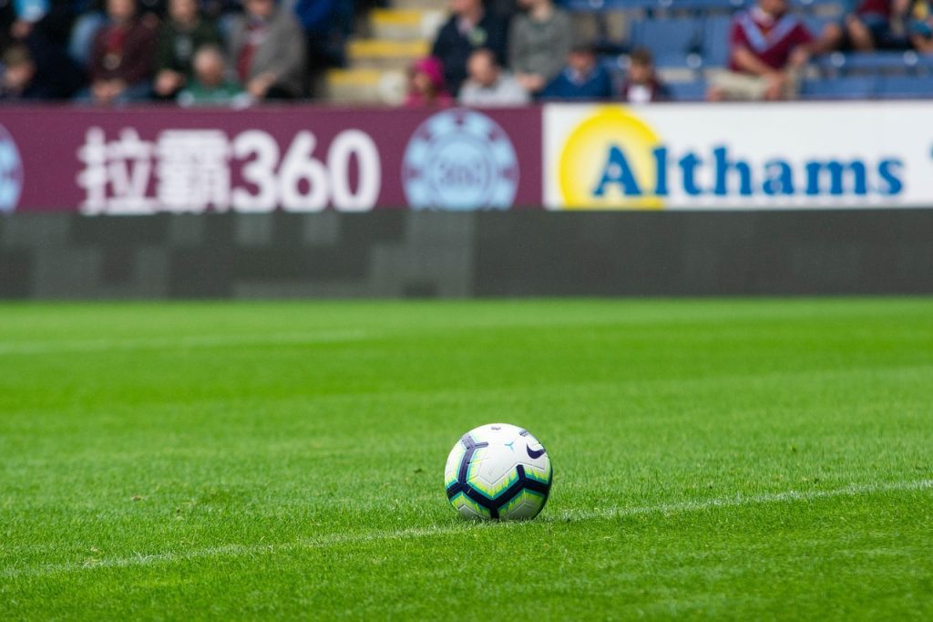 A white football with black and green accents rests on a green grass pitch, with a blurred background of stadium seating and advertising boards.
