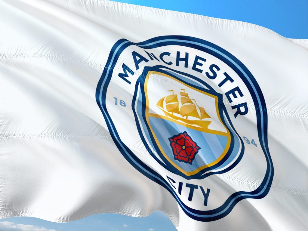 A white flag with the Manchester City Football Club logo waving in the wind. The logo is predominantly blue and white, featuring a ship, a red rose, and the words "MANCHESTER CITY" and "1894." This image is relevant to Premier League history.