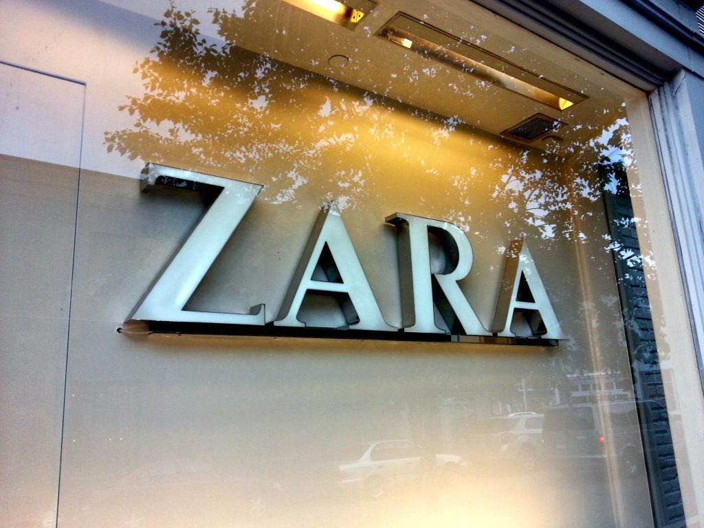 The silver "ZARA" brands name is visible on the exterior of a store, displayed on a clear glass window. Reflections of trees and the surrounding environment are visible on the glass, partially obscuring the view. The store's interior lighting is also reflected in the upper part of the window.