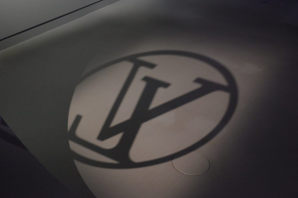 A shadow of the distinctive Louis Vuitton "LV" monogram brands logo is cast onto a light-colored surface, possibly a wall or floor. The interlocking letters 'L' and 'V' are enclosed within a circle, and a faint circular outline is visible to the lower right of the projected logo.