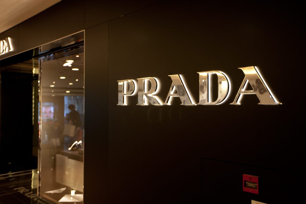 The silver, metallic "PRADA" brands name is prominently displayed on a dark, possibly black, storefront facade. The letters are three-dimensional and reflect light, indicating a polished surface. To the left, a glass entrance reflects the interior of the store, showing some merchandise and lighting.