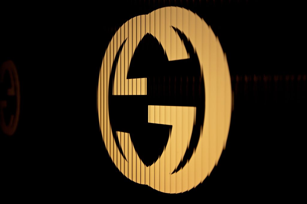 A glowing, golden Gucci brands logo, made of two interlocking Gs, is shown against a dark background, angled slightly to the left.