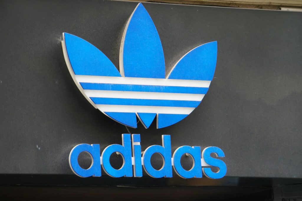 A close-up shot of an Adidas storefront sign featuring the blue and white Trefoil logo above the word "adidas" in lowercase blue lettering. This iconic emblem represents one of the world's leading sportswear brands.