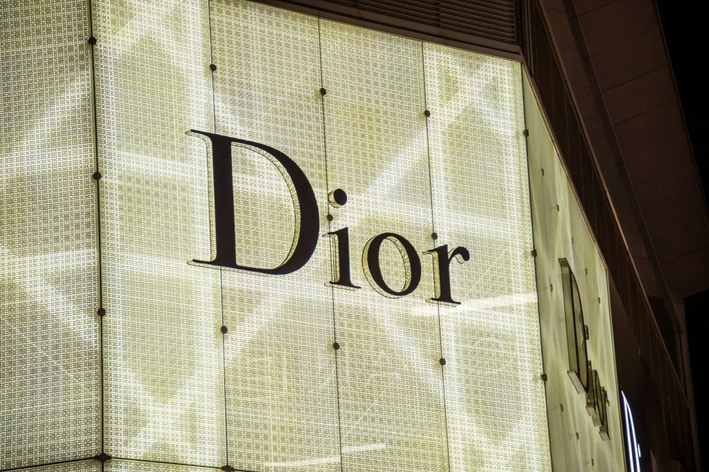 A brightly lit, large-scale Dior logo on the side of a luxury storefront. The black "Dior" text stands out against a textured, illuminated glass facade, showcasing one of the world's most prominent fashion brands.