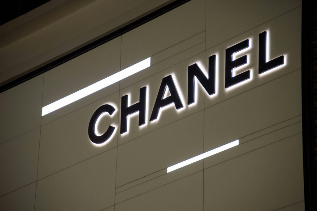 The prominent "CHANEL" brands name is displayed in black letters with a glowing white outline, set against a dark background. Two horizontal white light bars, one above and one below the text, frame the logo.