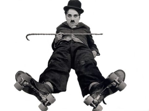 A grayscale image of Charlie Chaplin as his Tramp character, sitting with his legs splayed wide, wearing oversized roller skates. He has his signature bowler hat, small mustache, and holds his cane horizontally across his lap, looking directly at the viewer with a neutral expression. This iconic pose is from a classic comedy film.