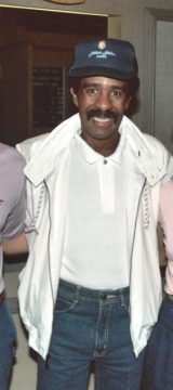 A smiling man, known for his comedy, wearing a navy blue baseball cap with a white emblem, a white polo shirt, a white jacket with a subtle pattern on the sleeves, and blue jeans. He has a mustache and short dark hair. He is looking directly at the camera.