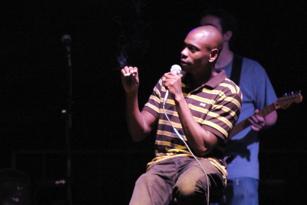 A male comedian, with a bald head, is seated on a stage, holding a microphone in one hand and a lit cigarette with smoke rising from it in the other. He is wearing a brown and yellow striped polo shirt and dark pants. In the blurred background, another person is visible holding a guitar, suggesting a band or musical accompaniment during the comedy act.