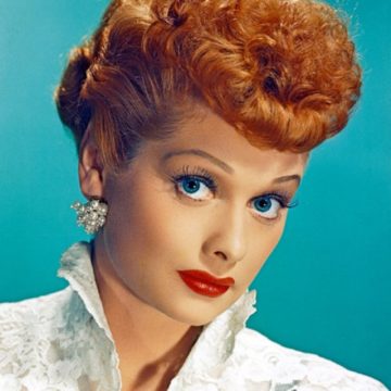 A headshot of a woman with bright red hair styled in an elaborate updo, vivid blue eyes, and prominent red lipstick, against a teal background. She is wearing a white blouse with a ruffled collar and sparkling earrings. Her expression is bright and engaging, typical of a comedic performer.