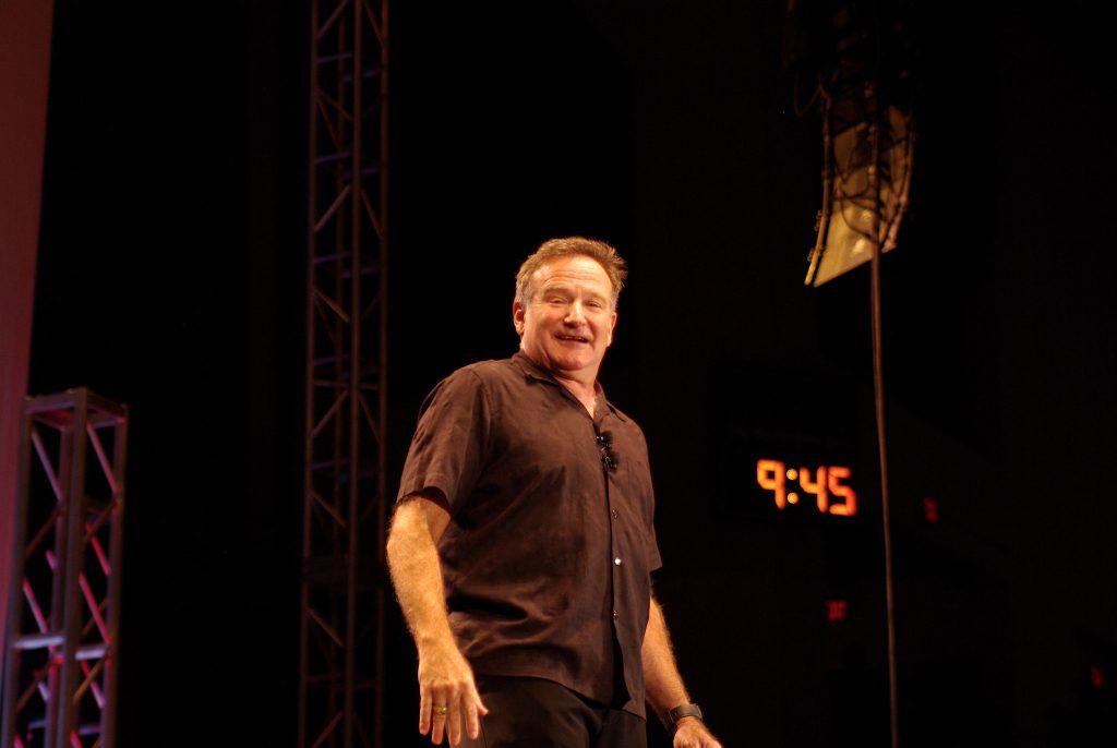 A smiling male comedian with light brown hair is standing on a dark stage, wearing a short-sleeved dark brown button-up shirt and dark pants. His hands are slightly bent at the wrist, resting in front of him. In the background, there's a red digital clock displaying "9:45" and stage rigging, typical of a comedy show or performance venue.