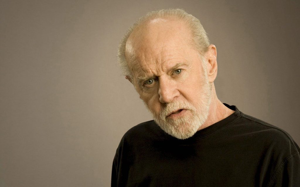 A close-up studio portrait of a male comedian with a bald head and a full, light-colored beard, looking directly at the camera with a quizzical or slightly confused expression. He is wearing a simple dark t-shirt against a plain gray background.
