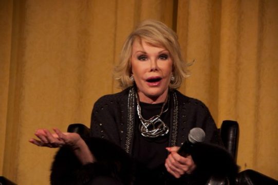 A female comedian with blonde hair, wearing a black top with several silver necklaces, is seated and holding a microphone in her left hand, while her right hand is open in a gesturing motion. She has a surprised or animated expression on her face, and a fur stole is draped over her lap. The background is a solid, warm golden-brown curtain, suggesting an indoor performance or interview setting. This image captures the essence of a comedic persona.