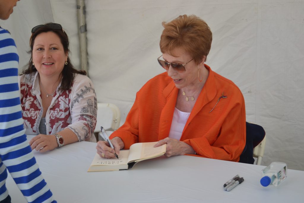 A woman with reddish-brown hair and sunglasses, wearing a bright orange blazer over a white top, is seated at a white table and actively signing a book with a pen. To her left, another woman with long dark hair, wearing a patterned blouse, smiles towards the camera. Part of a person in a blue and white striped shirt is visible on the far left, suggesting interaction with the people at the table. The setting appears to be outdoors under a white tent, possibly for a book signing event, though there's no direct indication of comedy.