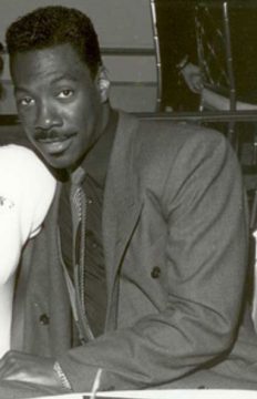 A black and white close-up of a man with dark, wavy hair and a mustache, wearing a suit and tie, looking towards the camera with a slight smile. The image captures him in a moment, potentially at an event, and he is widely known for his comedy work.