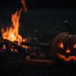 A carved Halloween jack-o'-lantern with a glowing face sits on the ground at night next to a crackling bonfire, symbolizing the holiday's ancient roots in the Celtic festival of Samhain where fires were lit for protection from spirits.