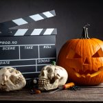 A film clapperboard rests next to a spooky carved jack-o'-lantern, two skulls, and candy corn, representing the theme of iconic Halloween horror movies.