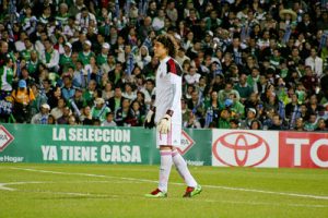 Guillermo Ochoa playing as goalkeeper for the Mexico National Team in a white kit during a match in a crowded stadium.