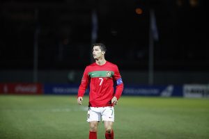 Cristiano Ronaldo standing on the pitch wearing the red Portugal long-sleeve home jersey with number 7 and a blue captain's armband.