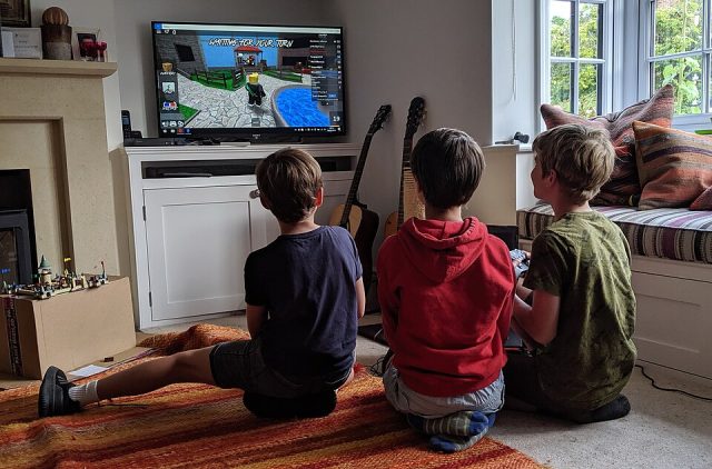 Three young boys sitting on a living room rug playing a Roblox game together on a large television screen.