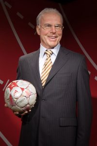 Franz Beckenbauer, one of football's legendary captains, wearing a dark pinstripe suit, glasses, and a striped tie, smiling while holding a Champions League football against a red background.