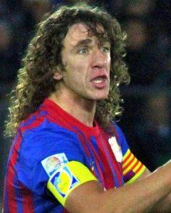Carles Puyol, one of football's legendary captains, wearing a blue and red Barcelona jersey and a yellow armband, looking intensely off-camera with his mouth open as if shouting instructions.