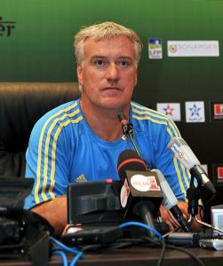 Didier Deschamps, one of football's legendary captains, seated at a press conference table with multiple microphones, wearing a light blue athletic shirt with yellow stripes.