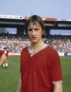 Johan Cruyff, one of football's legendary captains, standing on a pitch circa 1971 wearing a red jersey with white trim and a beaded necklace, with a stadium crowd in the background.