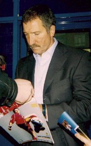 Graeme Souness, one of football's legendary captains, wearing a dark suit and light shirt, looking down as he signs a magazine for a fan.