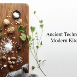 A minimalist flat-lay split between a wooden cutting board on the left and a clean white background on the right. The wooden board features a stone mortar and pestle, scattered coarse rock salt, fresh green herbs, whole almonds, and garlic cloves laid out for cooking. On the right side, elegant black text reads 'Ancient Techniques, Modern Kitchen'