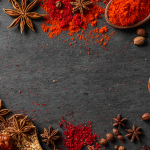 A dark, textured slate surface framed by a vibrant assortment of culinary spices, featuring scattered whole star anise pods, small brown seeds, and two rustic wooden spoons filled with bright red ground powder, leaving an empty space in the center for text.