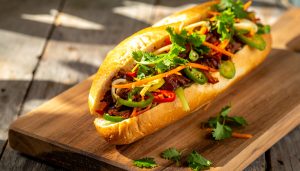 Vibrant food photography of a loaded Vietnamese bánh mì sandwich, a classic Southeast Asian street food, served on a wooden cutting board, featuring a crispy baguette packed with savory grilled meat, pickled carrots, daikon, sliced jalapeños, and fresh cilantro leaves under warm, natural sunlight.