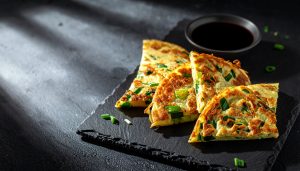Moody, dramatic food photography of crispy Taiwanese scallion pancakes, a popular night market street food, cut into wedges and served on a dark slate board with a side of dark soy dipping sauce.