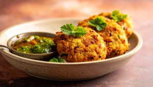 Appetite-inducing food photography of golden-brown Indian aloo tikki potato patties, a classic South Asian street food, arranged in a ceramic dish with a small bowl of vibrant green mint chutney, garnished with fresh cilantro leaves.