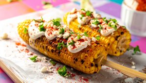 High-quality food photography of Mexican elote street food on wooden sticks, featuring charred sweet corn topped with creamy white sauce, crumbled cotija cheese, chili powder, and fresh cilantro.
