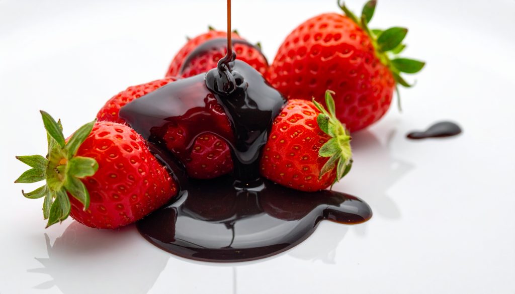 A close-up shot of glossy, dark balsamic vinegar glaze being poured over fresh red strawberries on a clean white surface, showcasing unique and unexpected flavor pairings.