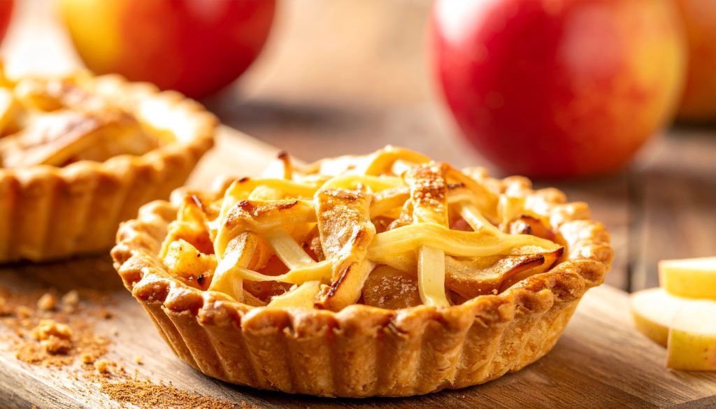 A golden, freshly baked mini apple tart with a lattice crust resting on a wooden board next to fresh red apples, representing traditional sweet and savory flavor pairings.