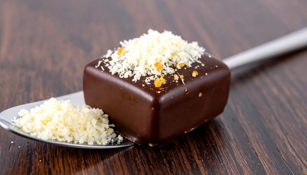 A gourmet dark chocolate praline topped with finely grated Parmesan cheese, presented on a silver spoon over a dark wooden surface to illustrate savory-sweet flavor pairings.