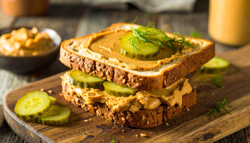 An appetizing, gourmet peanut butter and dill pickle sandwich on thick seeded bread, served on a rustic wooden board to showcase classic yet unusual flavor pairings.