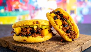 Vibrant food photography of two golden-brown Venezuelan arepas, a traditional South American street food, resting on a rustic wooden board, sliced open and packed with shredded beef, black beans, avocado, and yellow plantains against a colorful blurred background.