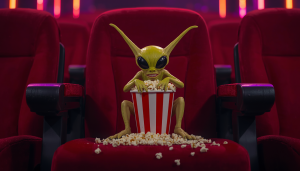A bizarre, green alien creature toy with large black eyes sitting in a red movie theater seat, holding a red-and-white striped popcorn bucket.