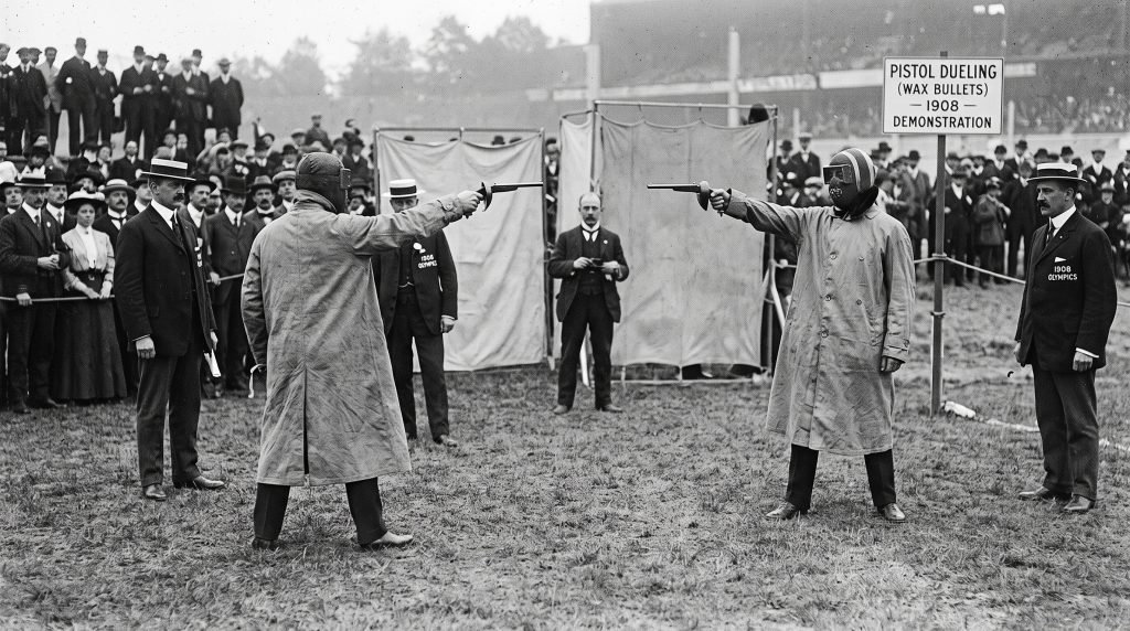 A black and white historic photograph of two men wearing heavy protective coats and face masks pointing pistols at each other for a pistol dueling olympic sport demonstration using wax bullets.
