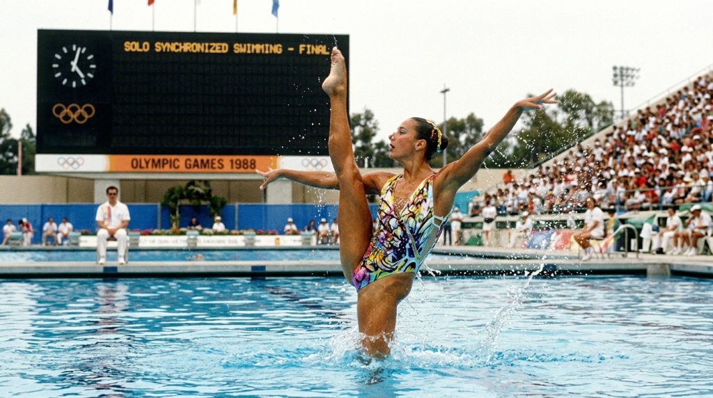 A vibrant color photograph of a female athlete performing the solo synchronized swimming olympic sport, executing a high leg split out of the water with a 1988 Olympic Games scoreboard in the background.